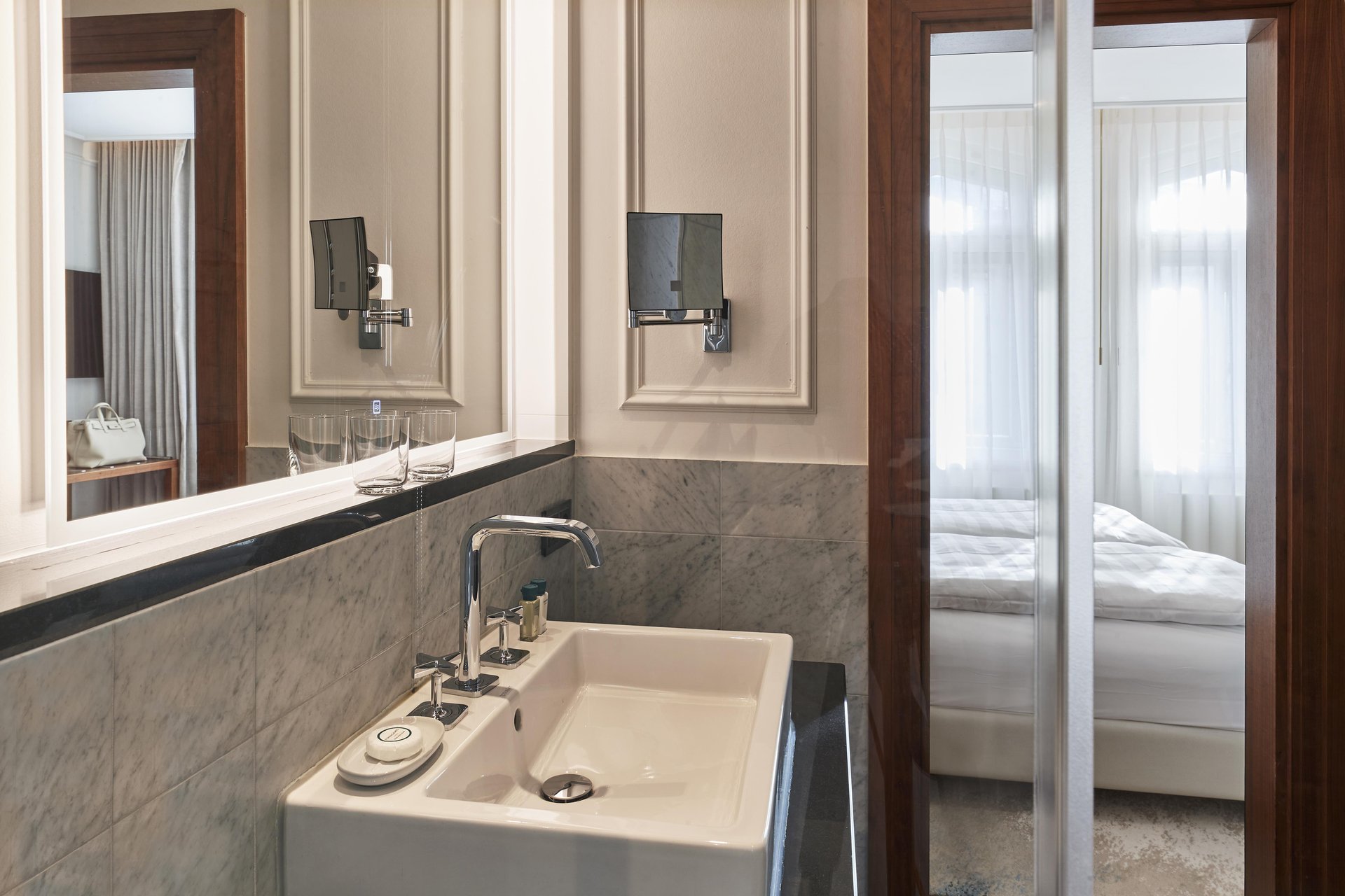 Marble bathroom vanity with chrome fixtures and view into bedroom at Hotel Vier Jahreszeiten Kempinski München
